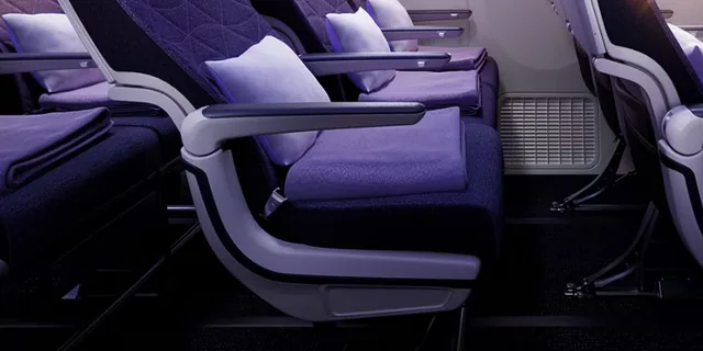 Kursi Economy Riyadh Air berbalut kain ungu, dengan bantal dan selimut tersedia di sandaran